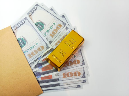 Gold bar placed on US dollar banknotes partially inside a brown envelope on a white background....