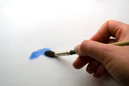 A woman's hand draws with blue paint. A hand with a brush draws on white paper with blue watercolor paint.の写真素材