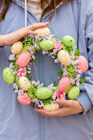 Homemade Easter wreath of bright eggs in women's hands on a blue background. Workshop on creating...
