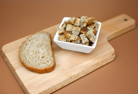 One piece of grain bread and croutons from it on a wooden board. Beige background. The concept of...