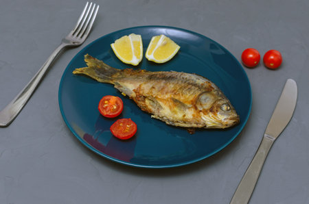Fried appetizing fish crucian carp with seasoning on a blue plate on a gray background decorated...