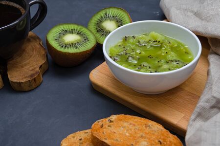 homemade kiwi jam in a white bowl close-up on a dark background with toast, morning coffee, fresh kiwi and a linen napkin. healthy eating concept. diet food.の写真素材