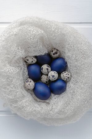Happy easter card - copy space. Blue easter and quail eggs in a snow-white woolen cozy nest on a white wooden background, selective focus image. bright contrast blank for holiday cardの写真素材
