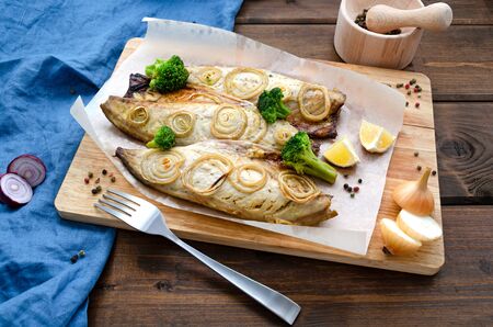 tender mackerel baked in the oven with chopped onion rings, broccoli and lemon on a dark wooden background with a blue napkin and fork. keto diet and healthy eating. copy spaceの写真素材