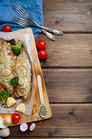 closeup tender baked mackerel in the oven with rings of chopped onion, broccoli, tomatoes and lemon on dark wooden background with blue napkin and forks. keto and flexitarian diet. copy spaceの写真素材