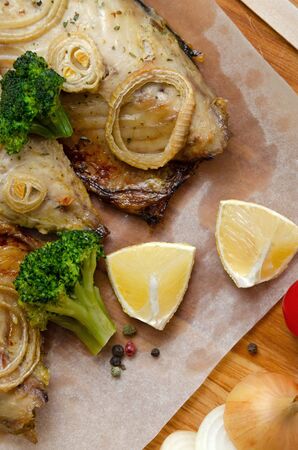 closeup fresh tender baked mackerel with rings of chopped onion, broccoli and lemon on a dark wooden cutting board in baking paper. keto and flexitarian diet and healthy eating. copy spaceの写真素材