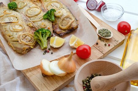 fresh baked tasty mackerel fish with lemon, sliced on filet with tomatoes, onion rings, pepper, herbs, olive oil and broccoli in baking paper on a cutting board on a white wooden background, close-upの写真素材