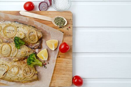 buttery tasty fish mackerel saba with lemon, sliced on filet with tomatoes, onion rings herbs and broccoli in baking paper on a cutting board on a white wooden background, close-up, copy spaceの写真素材