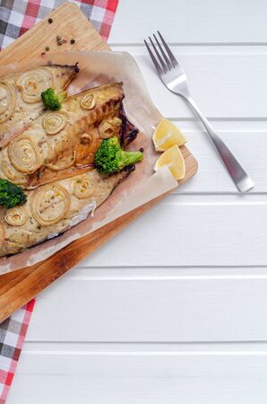 closeup tender baked mackerel in the oven with rings of chopped onion, broccoli, tomatoes and lemon on white wooden background with blue napkin and forks. keto and flexitarian diet. copy spaceの写真素材