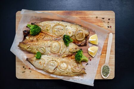closeup of fresh tender baked mackerel with chopped onion, broccoli and lemon rings on a black slate background on a wooden cutting board in baking paper. keto and flexitritis diet and healthy eating.の写真素材