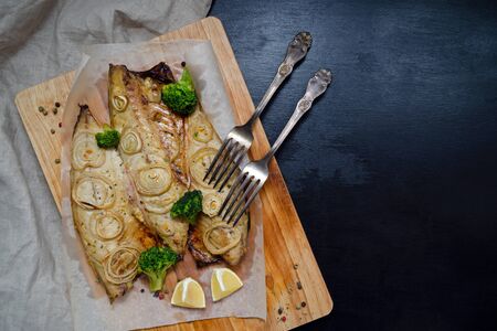 fresh tender baked mackerel with chopped onion, broccoli and lemon rings on a black slate background on a wooden cutting board in baking paper. keto and flexitritis diet and healthy eating. close upの写真素材