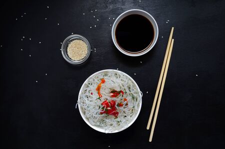 Asian glass fungoza rice noodles and bamboo sticks on a wooden board in a white round bowl with sesame seeds and soy sauce ready to eat on a black slate background with copy space.の写真素材