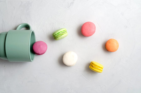 many multicolored crunchy delicious macarons dropped out of a ceramic cup on a gray concrete...