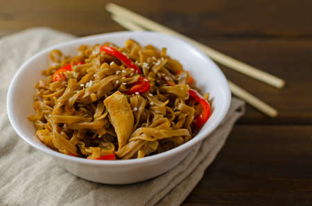 Appetizing asian japanese chinese cuisine with chicken and vegetables pepper with udon wok noodles in a white plate on a wooden table with wooden chopsticks, horizontal.の写真素材