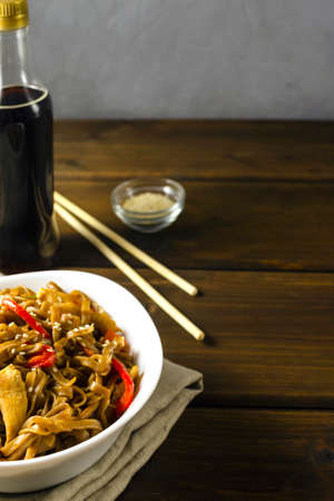 Appetizing asian japanese chinese cuisine with chicken and vegetables pepper with udon wok noodles in a white plate on a wooden table with wooden chopsticks, horizontal.の写真素材