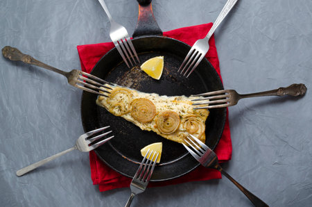 Fried baked mackerel trachurus with onions and dried herbs in an old frying pan in a towel, many different forks around. country style. Top view, gray concrete background, place for textの写真素材