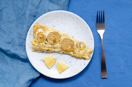 appetizing fish mackerel fillet piece on a plate baked cooked with onions and herbs with lemon slices on a blue background with a napkin and forkの写真素材