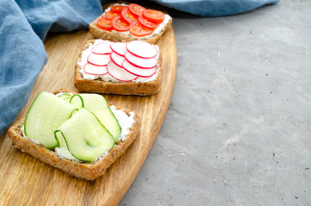 Vegetarian sandwiches with cream cheese, fresh cucumber, radish, tomatoes on a cutting wooden board with a towel on the concrete. background. healthy food diet weight loss fitnessの写真素材