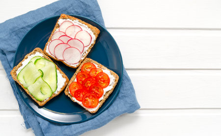 Assorted breakfast sandwiches with a slice of whole grain dark bread, cream cheese, cucumbers radishes cherry tomatoes on a white wooden background in blue plate. Top view. Flat lay. vegan diet postの写真素材