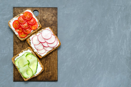Assorted breakfast sandwiches with a slice of whole grain dark bread cream cheese cucumbers radishes cherry tomatoes on a gray concrete background on a cutting board. Flat lay. vegan diet postの写真素材