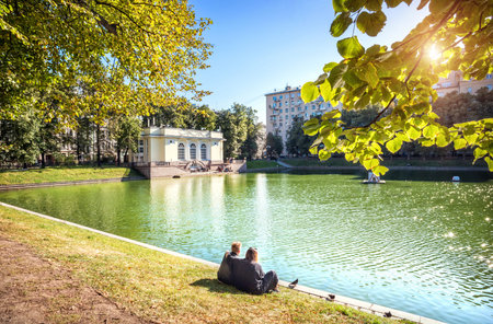 Pavilion on the Patriarch's Pond in Moscow, two lovers on the shore and the sun through the green of...