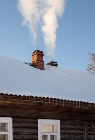 Wood-burning stove is blazing in a rustic log house. A column of white smoke rises from a brick chimney on a snow-covered roof.の写真素材