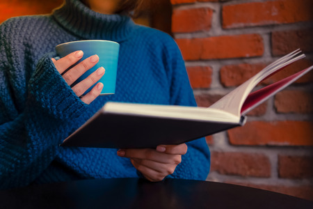 A girl in a beautiful blue sweater with a blue cup of coffee and a book in a cafe with brick walls and a wooden tableの写真素材