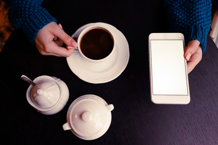 A girl in a blue sweater, with a perfect manicure in a cafe with coffee and turned on smartphone with isolated screen at the wooden table on a coffee breake.の写真素材