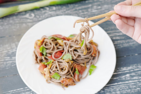 brown noodles or soba on chopsticks in girl's hand. fresh soba on the white plate with chicken and...