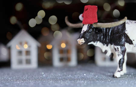 toy bull as symbol of 2021 year. happy new year concept. sparkling greeting card. the ox and toy houses with bokeh lights. cow in a santa's red hat. chinese zodiac character bull.の写真素材