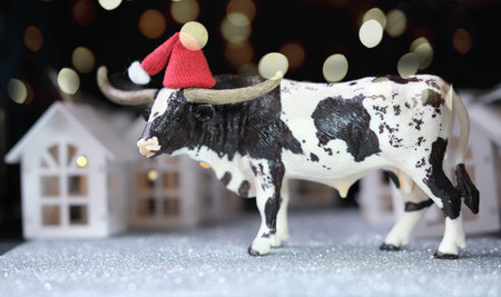 toy bull as symbol of 2021 year. happy new year concept. the ox and toy houses with bokeh lights. cow in a santa's red hat. chinese zodiac character bull.の写真素材