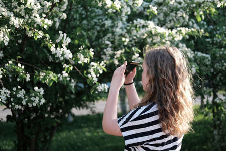 beautiful curly girl taking picture of blooming tree on a smartphone. teenager girl before a bush with white flowers. walking outdoors after quarantine. calm and peaceful place.の写真素材