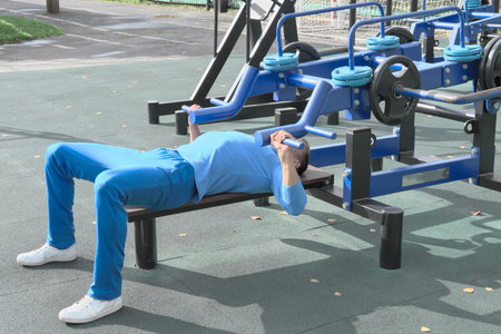 sporty slim man doing bench press using outdoors gym equipment. making an effort to lift a bar....