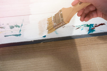 human hand holding brush painting white color on a white wooden table.