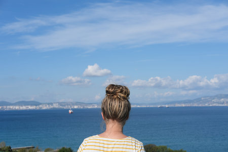 teenager girl with messy bun looking at the sea. sity and ship on the horizon. sea bay view. back view of a girl wearing t-shirt.の写真素材