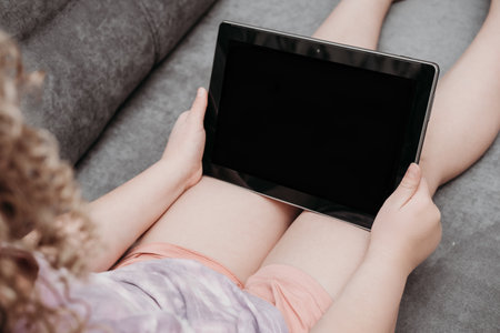 little girl holding the tablet pc. online education. distance learning. unrecognizable curly girl looking at tablet screen. mock up study. kid sitting on a sofa and watching online lesson.の写真素材