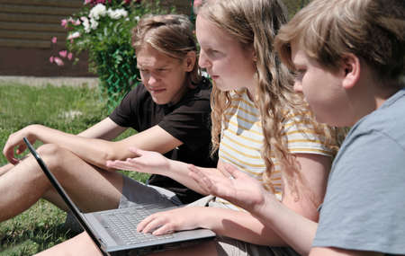 two teenager boys and a girl sitting on a grass and video chatting via laptop. communication and togetherness concept. emotional teens.の写真素材