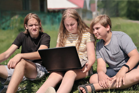 group of caucasian teenagers spending time on a lawn next to a house. friends looking at laptop and...