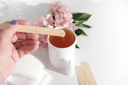 female hand holding a spatula with sugaring paste over white plastic container. depilation set next to a pink flowers on white background. beauty and skin care concept.の写真素材
