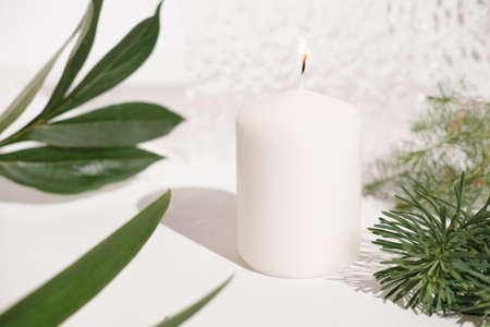 white candle and green leaves on white table with textured background. burning candle, minimal style home decor concept.の写真素材