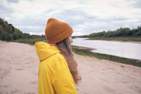 teenage girl wearing yellow raincoat and orange beanie standing on a river bank and looking aside. local travel concept. fall walking alone, teen autumn leisure.の写真素材