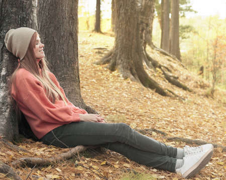 happy young red haired woman smiling and sitting under the tree. walking in an autumn forest. local travel. spending time in a park, relaxing and having funの写真素材