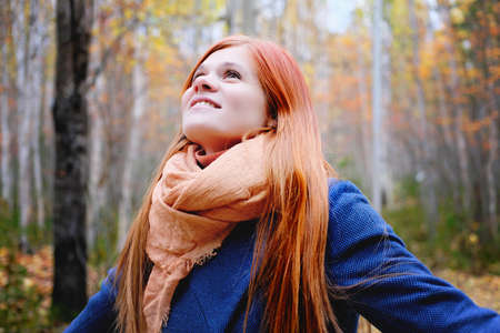 happy young red haired woman smiling and looking up. walking in an autumn forest. local travel. spending time in a forest, relaxing and having funの写真素材