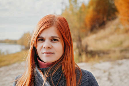 portrait of happy young red haired woman smiling and looking aside. local travel. spending time on a beach, relaxing and having fun. woman wearing blue coatの写真素材