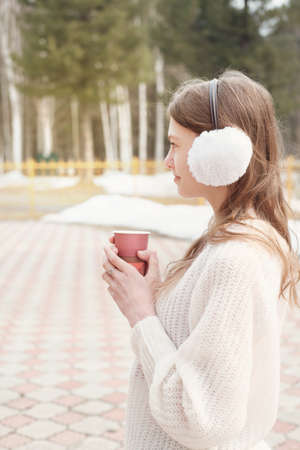 teenager girl holding a cup of coffee and looking aside while walking outdoors in winter. teen girl wearing earcuffs and white pullover. vertical shotの写真素材