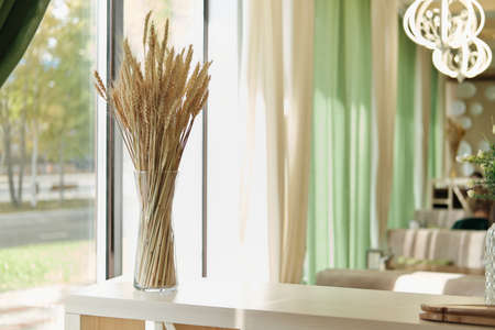 partial view of stylish interior of modern cafe, terrace with natural decor. ear of wheat in glass vase on light wooden shelf, green curtains. window view to a street. cozy place with minimal decorの写真素材
