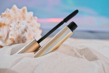 open eyelash mascara in golden tube on sandy beach next to a seashell. summer beauty and decorative cosmetic, make up tools. waterproof mascaraの写真素材