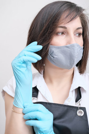 woman wearing apron putting on blue protective gloves. brunette woman with textile mask on a face...