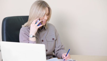woman talking with client and writing down details in a notebook. office space with laptop, copy...