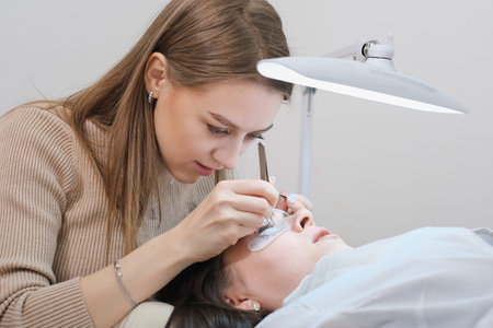 young woman beautician holding tweezers with false lash during lash extension procedure with client....